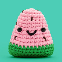 Sandy the Watermelon Crochet Kit by The Woobles features a cute crocheted watermelon plush with pink and green yarn, black bead eyes, and a smiling face—ideal for beginner crochet projects. Set against a teal background.