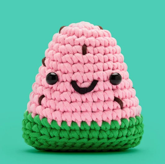Sandy the Watermelon Crochet Kit by The Woobles features a cute crocheted watermelon plush with pink and green yarn, black bead eyes, and a smiling face—ideal for beginner crochet projects. Set against a teal background.