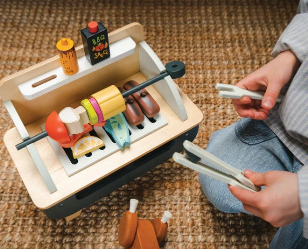 A child plays on a carpeted floor with the Tenderleaf Barbeque Play Set, which includes wooden skewers, fish, lemon slices, condiments, and tongs—ideal for tabletop play.