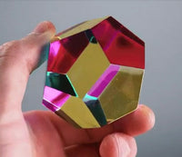 A hand holds The Mundus by CMY Cubes, a vibrant geometric crystal cube whose faceted surfaces reflect dazzling shades of pink, yellow, green, and blue for an engaging sensory experience.