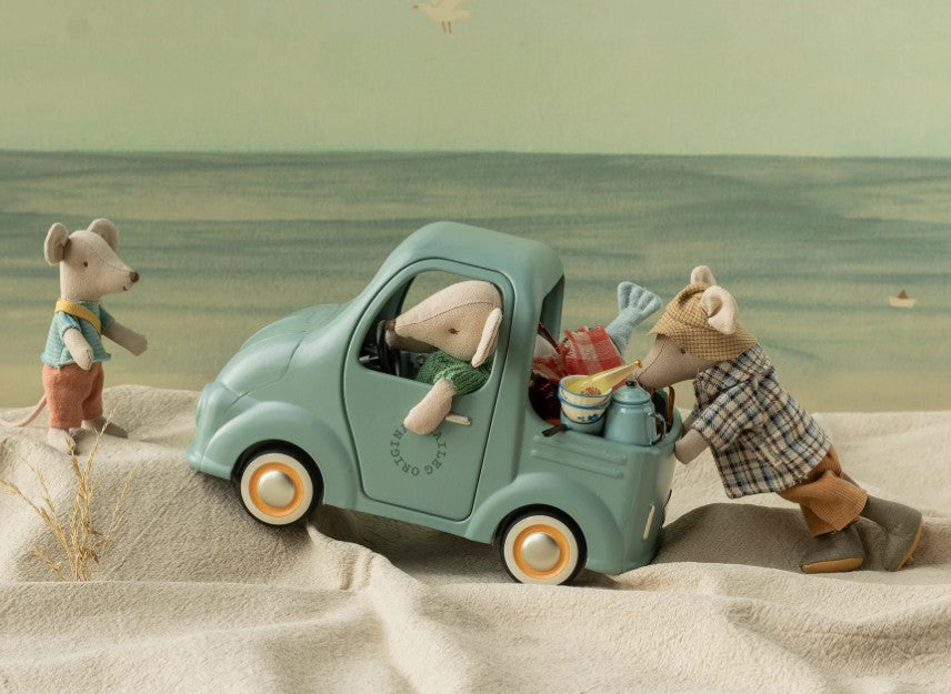 Three Maileg toy mice are on a sandy surface: one is driving the Maileg Truck, Mouse, another is pushing the miniature vehicle, and the third mouse stands nearby. The truck’s trunk is open and filled with small, mouse-sized travel accessories.