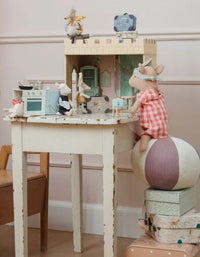 Several Maileg toy mice are gathered in a miniature kitchen scene, arranged around a small table and surrounded by charming accessories from the Maileg collection. One mouse wearing a pink dress sits atop a large ball, reaching toward the kitchen setup. Decorative boxes and a chair sit nearby, all set within the enchanting Maileg Castle hall, Mouse—capturing the whimsical atmosphere of this delightful creation by Maileg.