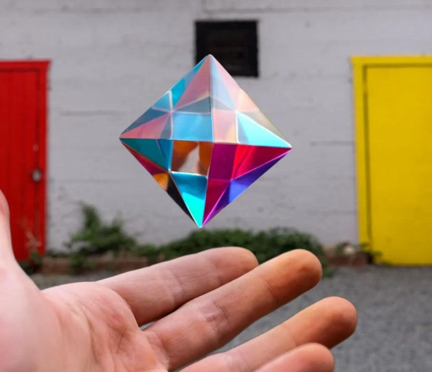 A hand is held under The Aether by CMY Cubes, a floating, reflective multicolored optical cube. Behind it are a white wall with a red door to the left and a yellow door to the right.