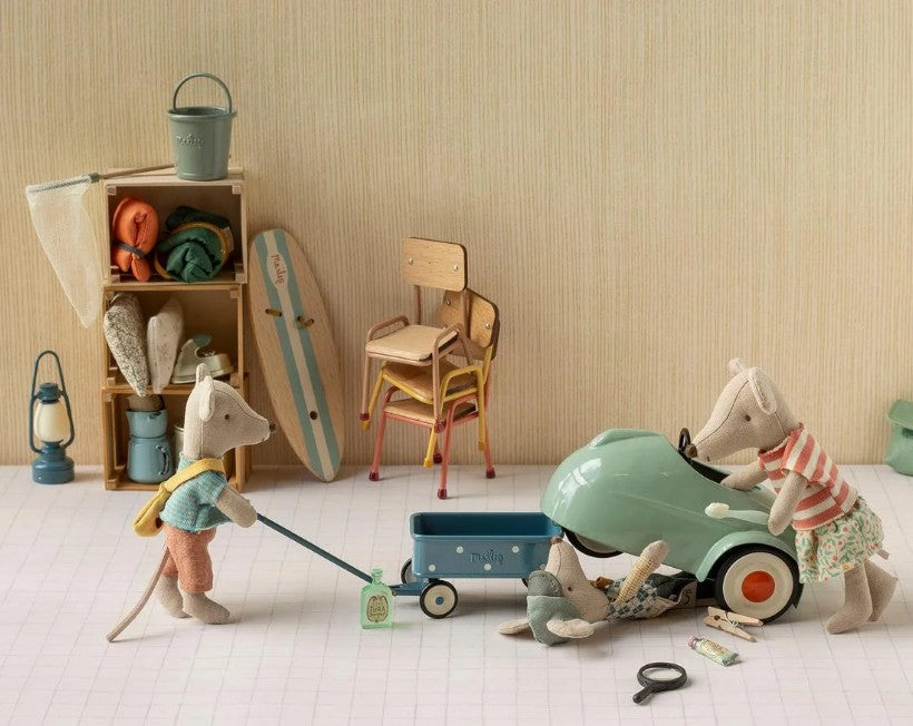 Three mouse toys dressed in charming outfits interact with the Maileg Car, Mouse in light mint, accompanied by a trailer and tiny pieces of luggage. Behind them, shelves display items such as a skateboard, chairs, a lantern, and blankets on a pale patterned floor.