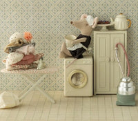 A toy mouse in a maid outfit sits on a washing machine holding a cup, while nearby a Maileg Iron and ironing board, Mouse by Maileg is surrounded by accessories such as a vacuum, teapot, cakes, and other charming details. Stuffed animals and delightful Maileg mice accessories complete the vintage dollhouse laundry room scene.