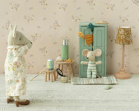 Two Maileg toy mice interact in a cozy room. One in a floral robe stands by food and accessories, while another in striped pajamas hangs from the Maileg Closet, Mouse - Soft Blue. A lamp and rug complete this charming dollhouse scene.