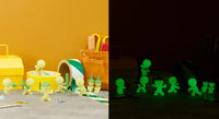 Several small yellow Smiski Construction Series figurines by Dreams USA are arranged near craft supplies in bright light on the left; on the right, the same scene is shown in darkness with the collectible blind-box figurines from Dreams USA glowing green.