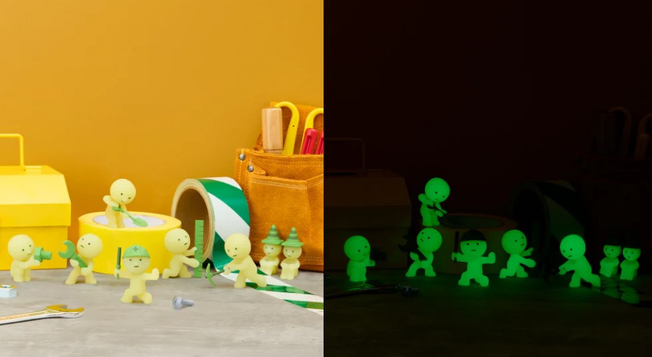 Several small yellow Smiski Construction Series figurines by Dreams USA are arranged near craft supplies in bright light on the left; on the right, the same scene is shown in darkness with the collectible blind-box figurines from Dreams USA glowing green.