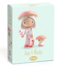 The DJECO Tinyly Amy & Mushy box features illustrations of Amy, a doll dressed in pink polka-dot pajamas and a pink mushroom hat, holding a pink suitcase beside her small mushroom companion. This collectible figure by DJECO captures woodland magic in every detail. Text on the box reads “Amy & Mushy” and “tinyly.”.