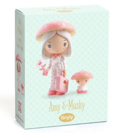 The DJECO Tinyly Amy & Mushy box features illustrations of Amy, a doll dressed in pink polka-dot pajamas and a pink mushroom hat, holding a pink suitcase beside her small mushroom companion. This collectible figure by DJECO captures woodland magic in every detail. Text on the box reads “Amy & Mushy” and “tinyly.”.