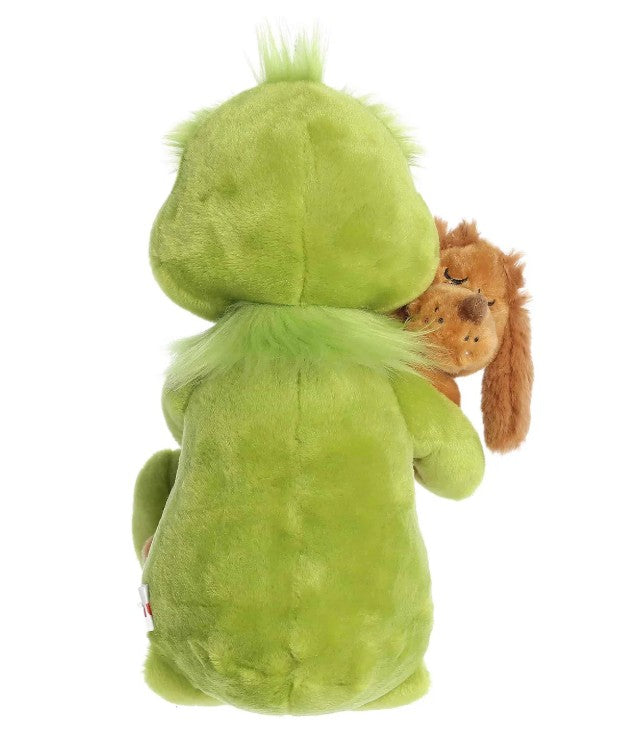 A green plush toy from the Aurora "Holiday Hugs" collection, pictured from behind, embraces a smaller brown dog plush. The cuddly toy boasts fuzzy fur and light green accents, while the brown dog features floppy ears and closed eyes. This heartwarming pair makes a perfect holiday gift for anyone who loves festive plush companions.