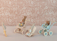 Three Maileg toy rabbits sit in the Maileg Stroller, Baby Mouse – Blue, role-playing as another rabbit looks on. The pink floral background adds charm to this imaginative play scene featuring the Maileg brand stroller.