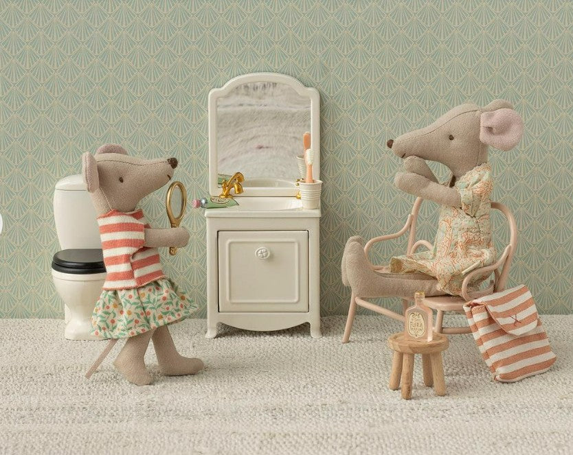 Two fabric Maileg mice are in a small bathroom with the Maileg Sink Dresser, Mouse - Off White. One holds a mirror in a striped top and floral skirt; the other sits by the vanity in pajamas. Also shown are a tiny stool, toilet, and striped bag.