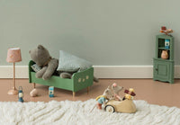 A plush bear rests on a small green bed with a checkered pillow, while nearby, toy mice zoom around in the cheerful Maileg Car, Mouse - Wheat by Maileg. Dollhouse furniture decorates the cozy, whimsical scene atop a fluffy white rug—making it perfect for imaginative play.