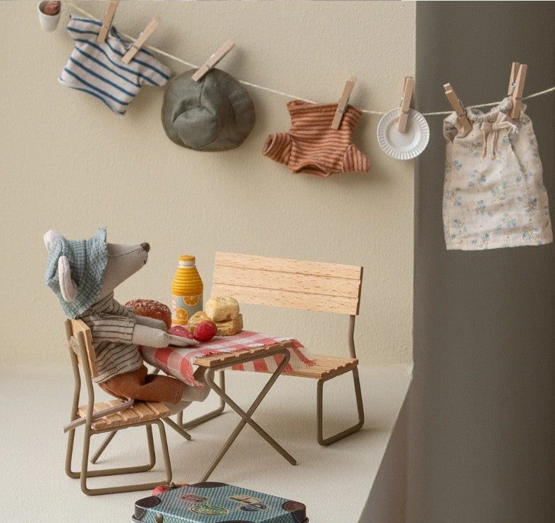The Maileg Hiker Mouse, Big Brother - Thin Stripes by Maileg sits at a tiny table with juice, bread, and an apple. Miniature clothes hang above, adding cozy charm to this whimsical camping-themed play scene.