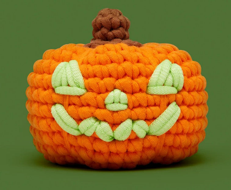 The Woobles’ Hal the Jack-o-Lantern Crochet Kit features a beginner-friendly orange pumpkin with a brown stem and green, smiling face, set against a solid green background.