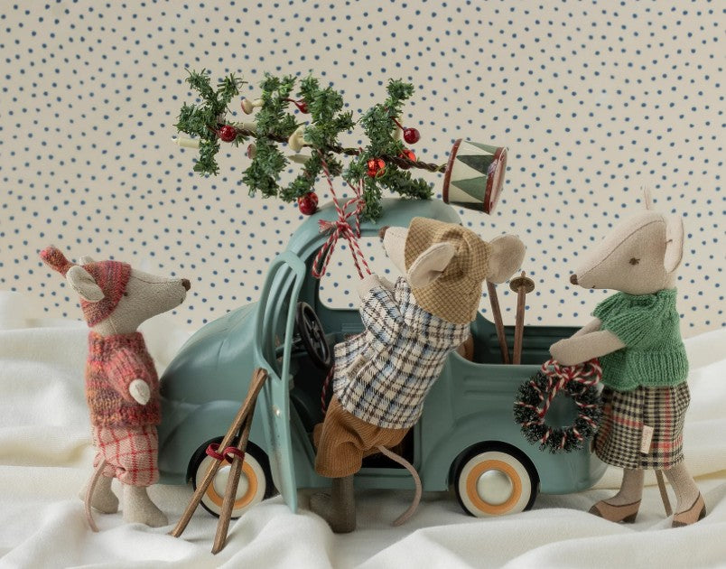 Three Maileg toy mice in festive winter outfits place a Christmas tree on the Maileg Truck, Mouse by Maileg. One mouse is dressed in plaid, one sports a green vest, and another wears a red hat. The mouse-size truck features a wreath on its front and rests atop fabric that resembles snow.