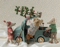Three Maileg toy mice in festive winter outfits place a Christmas tree on the Maileg Truck, Mouse by Maileg. One mouse is dressed in plaid, one sports a green vest, and another wears a red hat. The mouse-size truck features a wreath on its front and rests atop fabric that resembles snow.