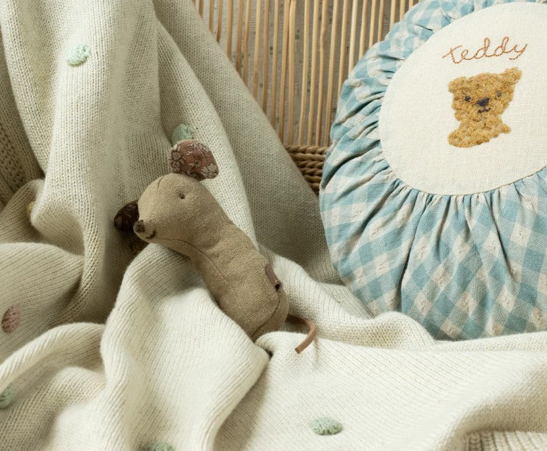 A Maileg Lullaby Friends Mouse Rattle in Light Umber rests on a cream knit blanket with pastel dots, next to a blue-and-white checkered cushion embroidered with a teddy bear—ideal for sensory play.