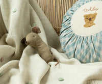 A Maileg Lullaby Friends Mouse Rattle in Light Umber rests on a cream knit blanket with pastel dots, next to a blue-and-white checkered cushion embroidered with a teddy bear—ideal for sensory play.