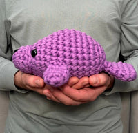 A person in a light gray shirt gently holds Woobles’ Phyllis the Manatee Extra Squeezy Crochet Kit, a handmade purple plush with black eyes—ideal for anyone seeking a beginner-friendly crochet project.