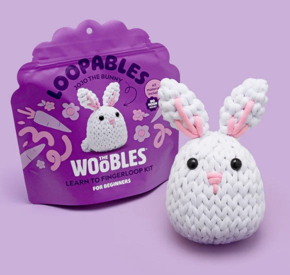 A small, crocheted white bunny with pink ears sits before a Jojo the Bunny Loopables Kit by The Woobles, labeled "Learn to Fingerloop Kit for Beginners." The background is light purple—perfect for fiber arts beginners.