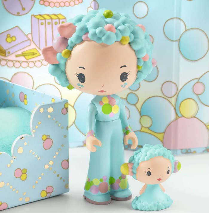 Two pastel-colored DJECO Tinyly figures with blue hair and floral accents from the DJECO Tinyly Blue Tinyroom stand on a decorated table, surrounded by a whimsical background featuring circles and book designs in soft colors—perfect for inspiring imaginative adventures in any miniature room playset.