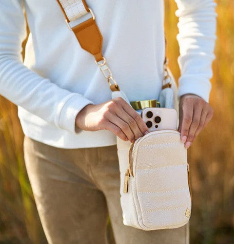A person in a white long-sleeve top and tan pants places a smartphone and tumbler into the compact Wader Full Brand HydroBag Handle Oatmeal Canvas outdoors, stylishly showcasing this versatile hydration bag accessory.