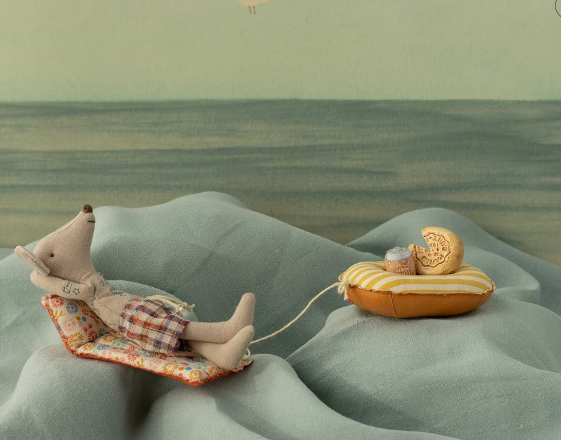 Maileg Beach Mouse With Air Mattress, Dad - Flower by Maileg features Dad Mouse lounging in plaid shorts on a patterned chair, with a string-attached yellow air mattress, creating a playful summer beach scene against a soft, fabric-like background.