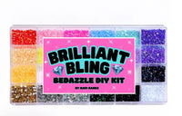 The Mavi Bandz Brilliant Bling Bedazzle Kit features a rectangular plastic container with 18 compartments of assorted colored rhinestones and a bright pink center label—ideal for creative rhinestone designs and DIY decorating.