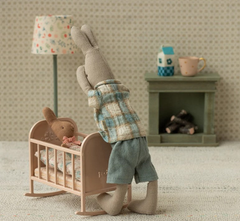 A Maileg stuffed rabbit in plaid kneels by a Maileg Cradle, My - Light Rose, with a smaller bunny inside. A toy lamp, fireplace, cup, and milk carton create a charming pretend play scene from Maileg.