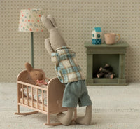 A Maileg stuffed rabbit in plaid kneels by a Maileg Cradle, My - Light Rose, with a smaller bunny inside. A toy lamp, fireplace, cup, and milk carton create a charming pretend play scene from Maileg.