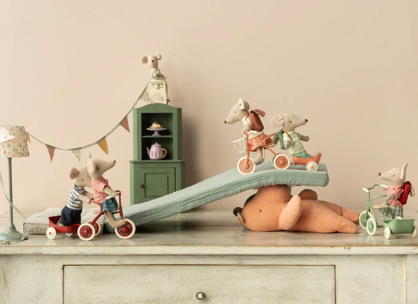 Stuffed toy mice on tricycles and scooters play on a cushion ramp atop a large teddy bear, with festive bunting and the Maileg Mouse Corner Cabinet in dark green by Maileg for miniature storage in the background.