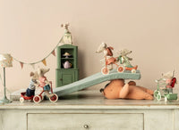 Stuffed toy mice on tricycles and scooters play on a cushion ramp atop a large teddy bear, with festive bunting and the Maileg Mouse Corner Cabinet in dark green by Maileg for miniature storage in the background.