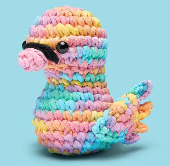 Tito the Swan Pastel Crochet Kit by The Woobles features thick pastel rainbow yarn and a beginner-friendly design. This small crocheted swan has black eyes, a pink beak, and colorful wings and tail, set against a light blue background.