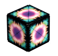 The Shashibo - Moon by Fun in Motion is a geometric cube, each face adorned with vibrant, psychedelic designs of purple, pink, green, and yellow rings inspired by celestial motifs, all set against a black background and encircled by pine tree silhouettes.