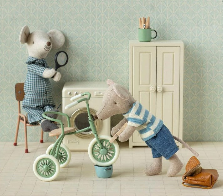 Two toy mice explore Maileg mouse accessories in a dollhouse laundry room; one in overalls examines clues with a magnifying glass, while the other checks miniature storage beside the Maileg Tricycle Garage (Green), washer, cabinet, mug, and shoes.