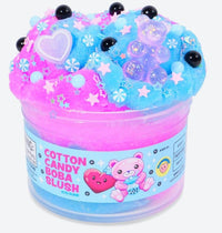 A clear jar labeled Cotton Candy Boba Slush Slime from Peachybbies Slime contains pink and blue fluffy slime with a delightful slushy texture, topped with beads, stars, and candy-shaped charms. The label features cute cartoon bears and hearts, making this sensory slime both adorable and fun to play with.