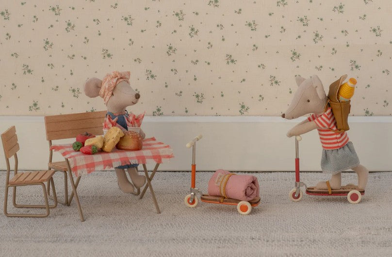 Two Maileg Mice create a charming scene: one sits at a table with food, while the other rides the Maileg Kick Board, Mouse - Orange by Maileg, carrying a blanket and basket. Light floral wallpaper decorates the background.