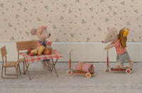Two Maileg Mice create a charming scene: one sits at a table with food, while the other rides the Maileg Kick Board, Mouse - Orange by Maileg, carrying a blanket and basket. Light floral wallpaper decorates the background.