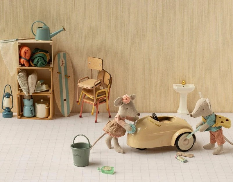 Two Maileg mice wash the Maileg Car, Mouse - Wheat in a cozy room. Behind them are chairs, a cupboard with towels and household items, and a small sink. The scene, styled in soft, neutral colors by Maileg, invites imaginative play.